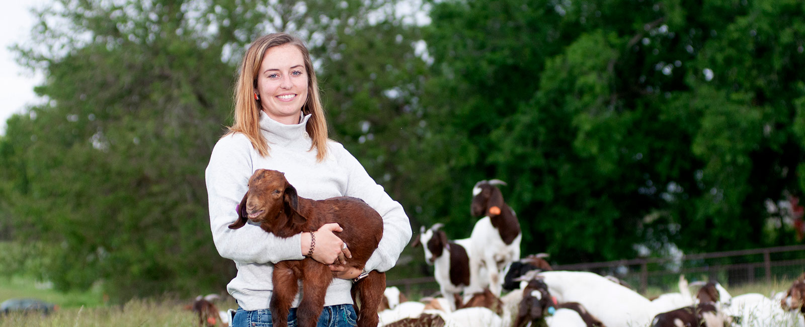 Instructor Alison Crane holds a young goat in a field while other goats gather behind her.